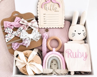 May include: A baby gift box filled with pink and white items. Includes a plush bunny, a rainbow-shaped teether, a set of headbands with bows, a wooden sign that says "hello world", and a personalized blanket embroidered with the name "Ruby".