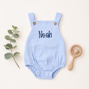 Könnte beinhalten: Hellblaue Baby-Latzhose mit dem Namen "Noah" in Marineblau gestickt. Die Latzhose hat Holzknöpfe und einen Druckknopfverschluss. Eine Holzrassel und ein Zweig mit Grünzeug sind ebenfalls im Bild.