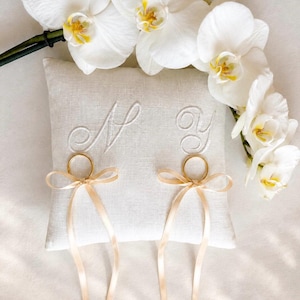May include: A square, off-white linen ring bearer pillow featuring embroidered cursive letters M and Y. Two gold-toned metal rings are attached to the pillow, each secured with a light beige satin ribbon tied into a bow with long, trailing ends.