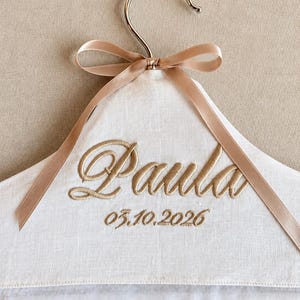 May include: A white fabric-covered clothes hanger featuring a metallic hook, a beige ribbon bow, and gold embroidery reading "Paula" and "03.10.2026". The bottom edge of the hanger is finished with a delicate white scalloped lace trim detail.