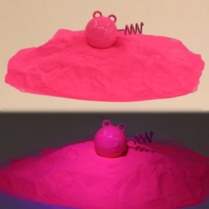 Puede incluir: Una pelota redonda de color rosa con un resorte unido a ella, sentada sobre un montón de arena rosa. La pelota y la arena brillan intensamente bajo una luz negra.