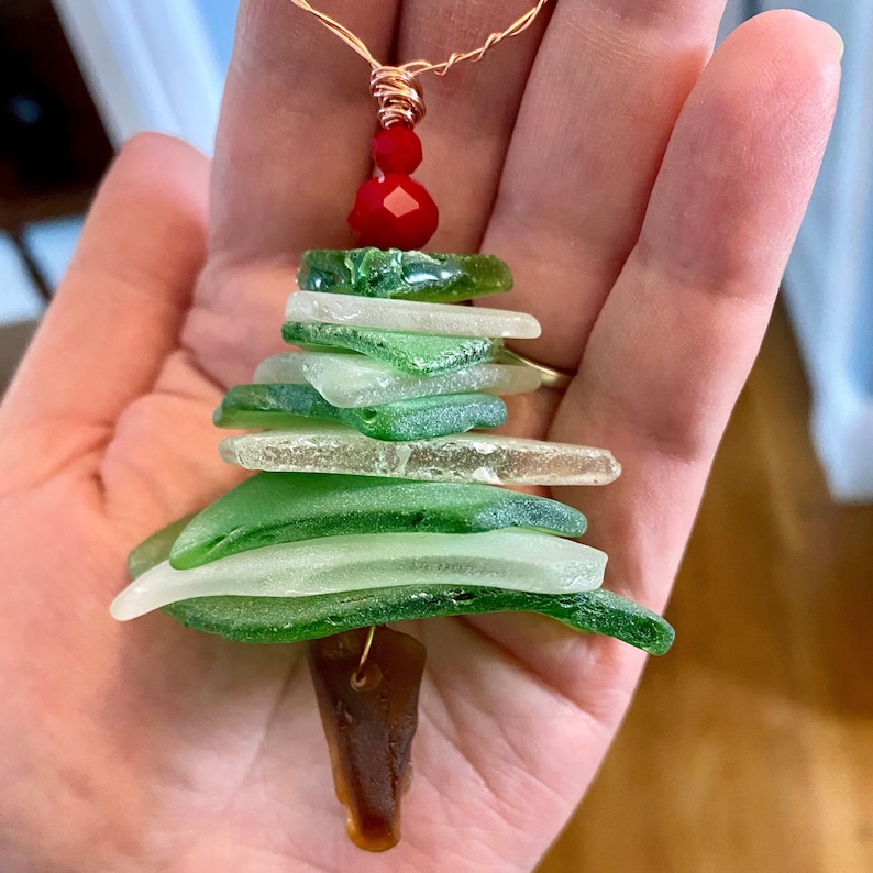 May include: A Christmas tree ornament made of layered green and white glass pieces. The ornament is topped with a red bead and has a brown glass base.