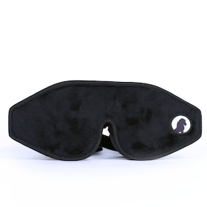 May include: Black sleep mask with a soft, velvety texture. The mask has a contoured shape to fit around the eyes and a black elastic strap. A small, circular patch with a silhouette is on the right side.
