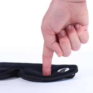 Puede incluir: Una m&aacute;scara para dormir negra con una textura suave y afelpada. La m&aacute;scara tiene una forma curvada para adaptarse a los contornos de la cara y un logotipo blanco en el lateral. Un dedo presiona la m&aacute;scara, destacando su suavidad.