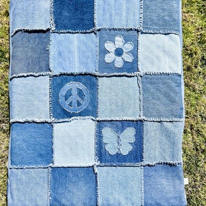 May include: A rectangular patchwork quilt made of various shades of blue denim fabric squares with frayed edges. The design features appliquéd denim shapes including a six-petal flower, a peace sign, and a butterfly centered on specific squares.