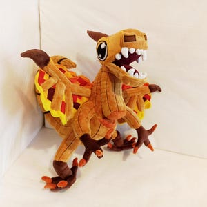 May include: A plush toy of a brown and orange dinosaur-like creature with large white teeth and a wide open mouth. The toy has large eyes and wings with yellow, red, and orange accents. The toy is made of soft fabric.