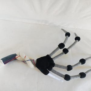 May include: A plush, articulated hand with a skeletal design. The fingers are gray with black accents, and the wrist area features a mix of white, blue, and red fabric. The palm is black, and the wrist is white. This item is likely a costume accessory.