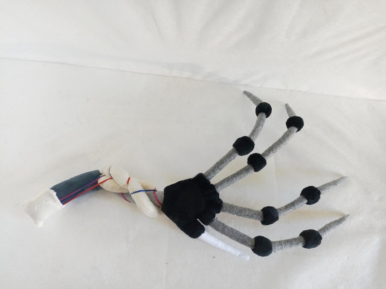 May include: A plush, articulated hand with a skeletal design. The fingers are gray with black accents, and the wrist area features a mix of white, blue, and red fabric. The palm is black, and the wrist is white. This item is likely a costume accessory.