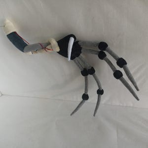 May include: A handmade, gray and black felt spider leg with a white and blue arm section. The spider leg has black joints and a needle extending from the arm. The felt spider leg is likely a Halloween or costume accessory.