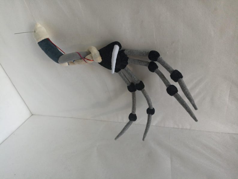 May include: A handmade, gray and black felt spider leg with a white and blue arm section. The spider leg has black joints and a needle extending from the arm. The felt spider leg is likely a Halloween or costume accessory.