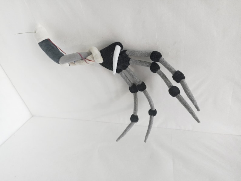 May include: A gray and black felt articulated hand with a white cuff and a needle attached. The hand has gray fingers with black joints. The wrist is gray and black. The hand is set against a white background.