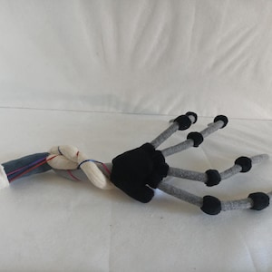 May include: A plush, stylized creature arm with a black hand and gray fingers ending in black spheres. The arm transitions to a twisted, gray and white section with blue and red accents, and a white cuff.
