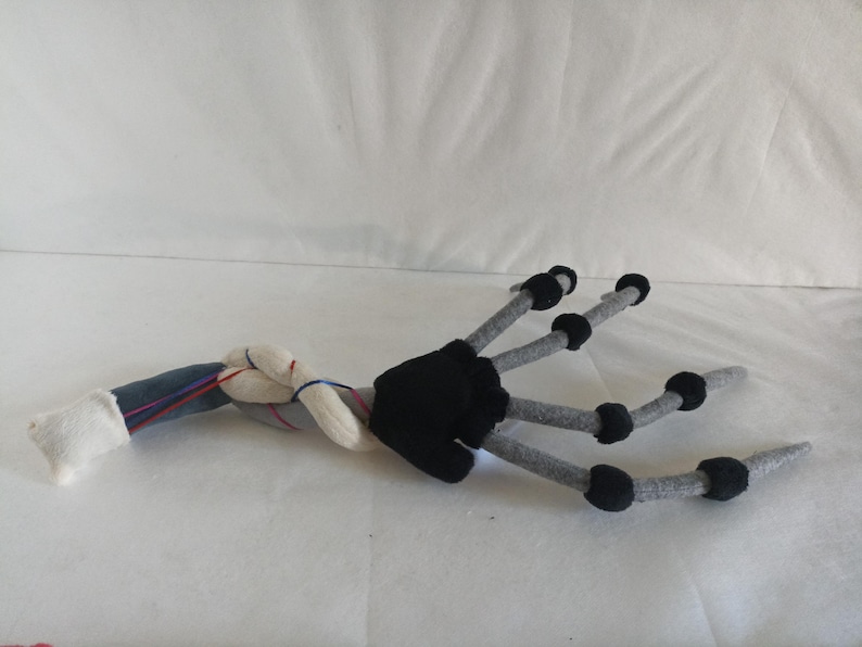 May include: A plush, stylized creature arm with a black hand and gray fingers ending in black spheres. The arm transitions to a twisted, gray and white section with blue and red accents, and a white cuff.