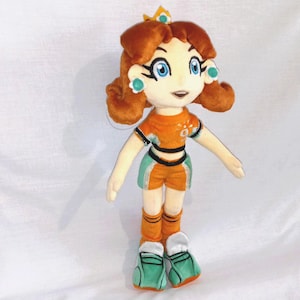 May include: A plush doll of a character with orange hair, blue eyes, and a small crown. The doll is wearing an orange and green sports uniform with matching shoes. The doll has a smiling expression and is standing against a white background.