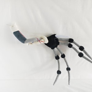 May include: A handmade, plush, articulated creature with a segmented body. The creature features a black torso, gray limbs with black joints, and a denim-like arm. The design is whimsical and appears to be a unique art piece.