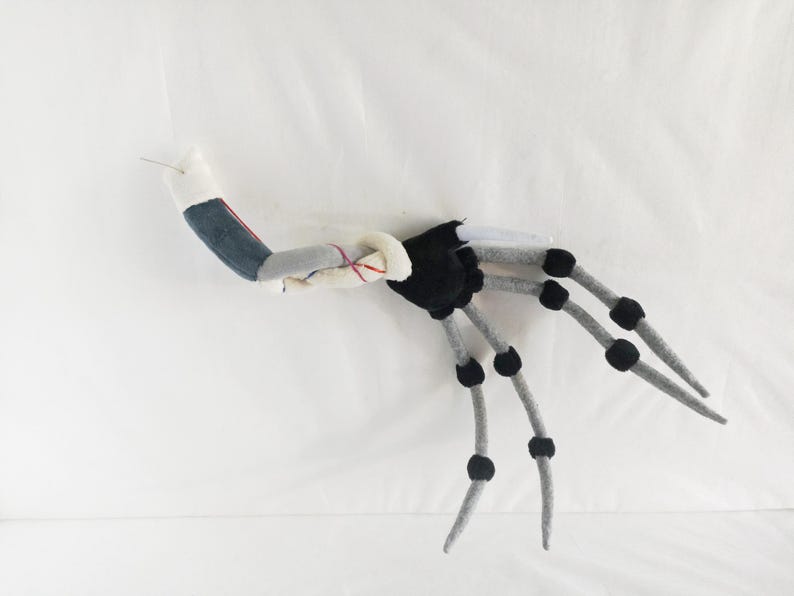 May include: A handmade, plush, articulated creature with a segmented body. The creature features a black torso, gray limbs with black joints, and a denim-like arm. The design is whimsical and appears to be a unique art piece.