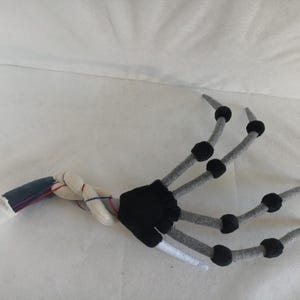 May include: A plush, articulated hand with a skeletal design. The fingers are gray with black joints, and the wrist area is a combination of white, blue, and red fabric. The palm is black, and the arm is white.