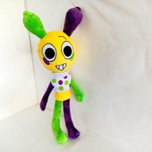 Peut inclure: Une peluche colorée avec une tête jaune, des oreilles vertes et violettes, et de grands yeux noirs. Le jouet a une chemise blanche à pois verts et violets, et des membres verts et violets. La peluche est sur fond blanc.