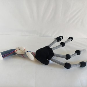 May include: A plush, stylized hand with gray fingers and black accents. The wrist area features a denim-like fabric, white fabric, and red and blue threads. The hand is lying on a white surface.