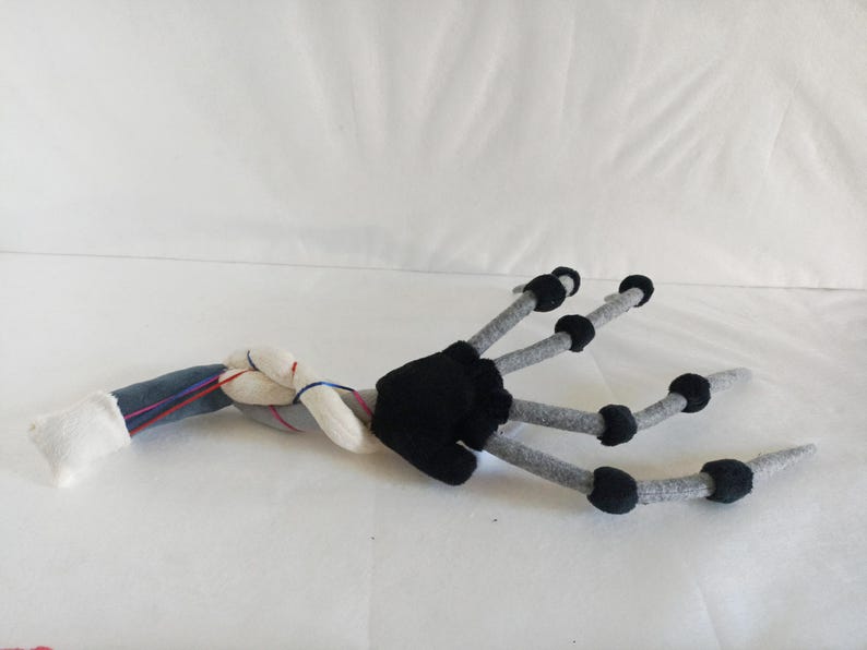 May include: A plush, stylized hand with gray fingers and black accents. The wrist area features a denim-like fabric, white fabric, and red and blue threads. The hand is lying on a white surface.