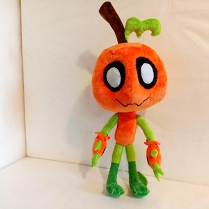 May include: A plush toy in the shape of a pumpkin-headed character. The toy is primarily orange with green accents on the arms, legs, and top of the head. It has large white eyes with black outlines and a stitched smile.