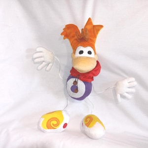 May include: A plush toy of a cartoon character with orange hair, a red scarf, and a purple body. The character has white hands and feet, and the feet are yellow with orange swirls. The toy is approximately 12 inches tall.