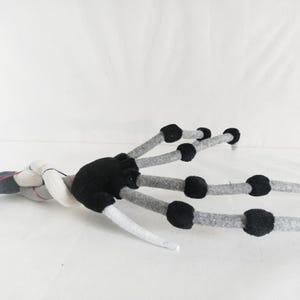 May include: A handmade felt claw with a black palm and gray fingers. The fingers have black circular accents and end in white tips. The claw is lying on a white surface.