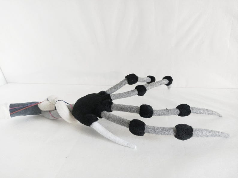 May include: A handmade felt claw with a black palm and gray fingers. The fingers have black circular accents and end in white tips. The claw is lying on a white surface.