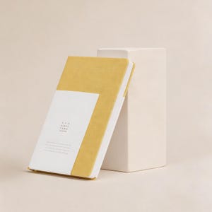 Puede incluir: Un cuaderno amarillo mostaza con una etiqueta rectangular blanca, apoyado contra un bloque rectangular blanco. La cubierta del cuaderno es de color liso y la etiqueta presenta texto. Las páginas del cuaderno son blancas.