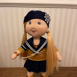 Könnte beinhalten: Eine handgemachte Puppe mit blonden Haaren, die ein blaues und weißes Matrosen-Outfit, eine braune Baskenmütze mit einem weißen Rad-Design und braune Schuhe trägt.
