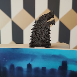 Peut inclure: Un marque-page en plastique noir imprimé en 3D en forme de tête de loup, un symbole populaire de la série télévisée Game of Thrones. Le marque-page est posé sur un livre avec une ville en bleu et gris sur la couverture.