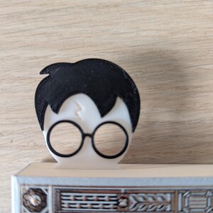Peut inclure: Un marque-page imprimé en 3D en noir et blanc représentant un dessin animé d'Harry Potter avec sa cicatrice en forme d'éclair et ses lunettes rondes.