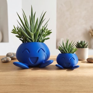 May include: Two vibrant blue planters shaped like smiling, round characters with small feet, each containing a spiky green succulent plant. The planters feature carved, happy facial expressions with curved eyes and an open mouth design.