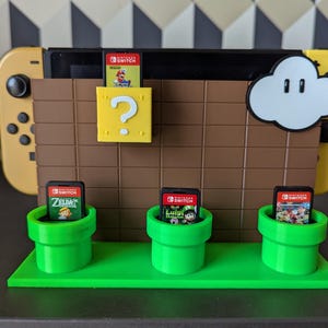 Puede incluir: Un soporte de cartuchos de juego de Nintendo Switch impreso en 3D de color verde y marrón con un bloque de interrogación, una nube y tres cartuchos de juego. Los cartuchos son para los juegos Zelda, Luigi's Mansion 3 y Super Mario Odyssey.