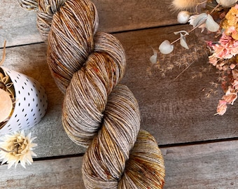 May include: A skein of hand-dyed yarn in shades of brown, gold, and cream. The yarn is arranged in a twisted, looped shape, showcasing the color variations. Dried flowers and a decorative white container are in the background, suggesting a craft or home decor theme.