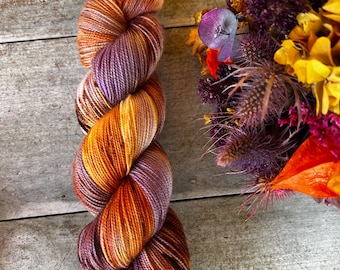 May include: A skein of yarn with shades of purple, orange, and yellow. The yarn is twisted into a spiral shape and is lying on a wooden surface.