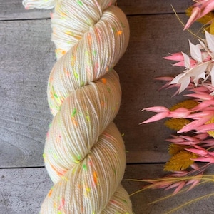 Puede incluir: Un ovillo de hilo blanco con motas de color verde neón, naranja y rosa. El hilo está dispuesto en una bobina suelta.