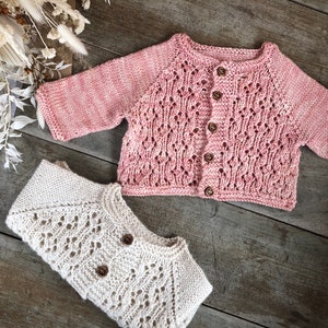 Puede incluir: Dos cárdigans de punto para bebé, uno rosa y otro blanco, con un delicado patrón de encaje y cierres de botón.