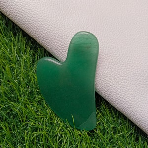 Op de afbeelding: Een hartvormig groen jade gua sha-gereedschap, een traditioneel Chinees massagegereedschap, op een groene gras achtergrond.