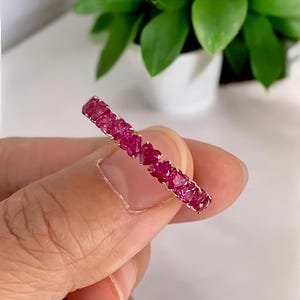 May include: A gold ring set with numerous heart-shaped, vibrant pink gemstones. The ring is held up against a blurred background of green foliage and a white surface, showcasing the ring's design and color.