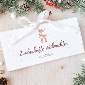 Könnte beinhalten: Eine weiße Geschenkbox, gebunden mit einem weißen Band, verziert mit einem Cartoon-Rentier mit Hut und Schal. Auf der Box steht "Zauberhafte Weihnachten" und der Name "ALEXANDER". Festliche Weihnachtsdekoration umgibt die Box.