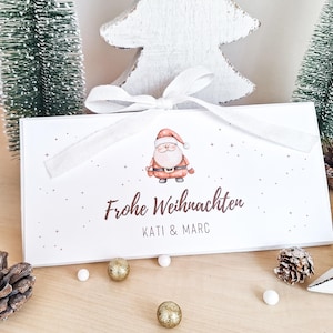 Könnte beinhalten: Eine weiße Geschenkbox mit einer Illustration des Weihnachtsmanns und dem Schriftzug "Frohe Weihnachten" in Braun. Die Box ist mit einem weißen Band gebunden und mit kleinen Sternen verziert. Weihnachtliches Thema.