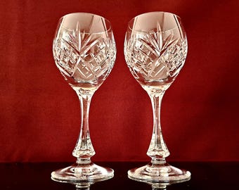 May include: Two clear crystal wine glasses with intricate diamond and starburst patterns. The glasses have long stems and round bases, set against a deep red background. The cut crystal design adds a touch of elegance.