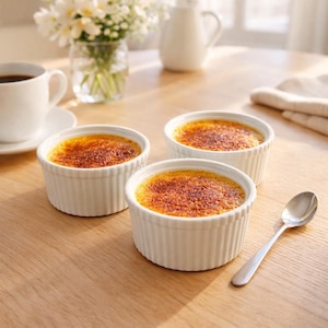 May include: Three white ramekins filled with crème brûlée, featuring a caramelized sugar crust. A silver spoon rests on the wooden table next to the desserts. A cup of coffee and a vase of flowers are in the background.