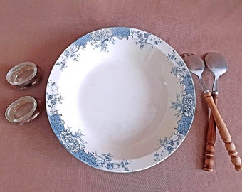 May include: A white ceramic bowl with a blue floral pattern around the rim. The bowl is empty and sitting on a brown surface. There are two small glass containers and two wooden handled spoons next to the bowl.