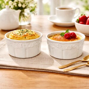 May include: Two white, decorative ramekins filled with food, one with a golden-brown top and the other with a yellow top. The ramekins sit on a beige placemat, with gold spoons, a cup of tea, and a bowl of strawberries in the background.