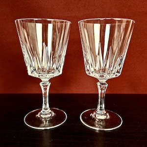 Pode incluir: Dois copos de vinho de cristal transparente com um design detalhado em corte. Os copos têm uma taça alargada, uma haste fina e uma base redonda. O fundo é vermelho escuro e os copos estão sobre uma superfície escura.