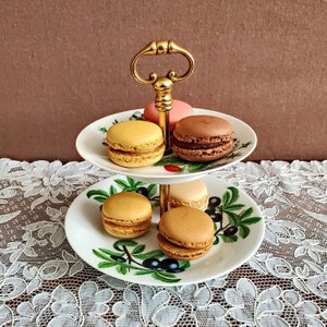 Peut inclure: Un présentoir à gâteaux à deux niveaux avec une poignée dorée, contenant plusieurs macarons colorés. Les assiettes blanches sont ornées d'un motif de branches d'olivier vertes. Les macarons sont de couleurs marron, beige et rose.