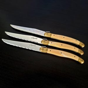 May include: Three Laguiole steak knives with serrated blades and cream-colored handles. The blades are silver, and the handles have brass accents. The knives are arranged on a dark surface.