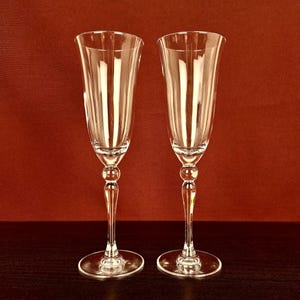 Può includere: Due flûte da champagne trasparenti con stelo decorativo e coppa ampia. Le flûte sono in vetro e presentano linee verticali incise nella coppa. Le flûte sono su una superficie marrone scuro con sfondo rosso.